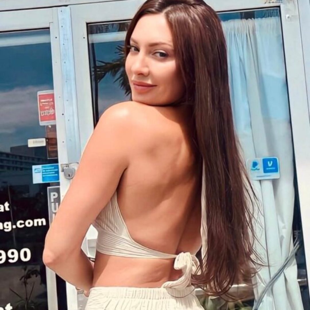 A woman with long brown hair poses outdoors in a backless white top.